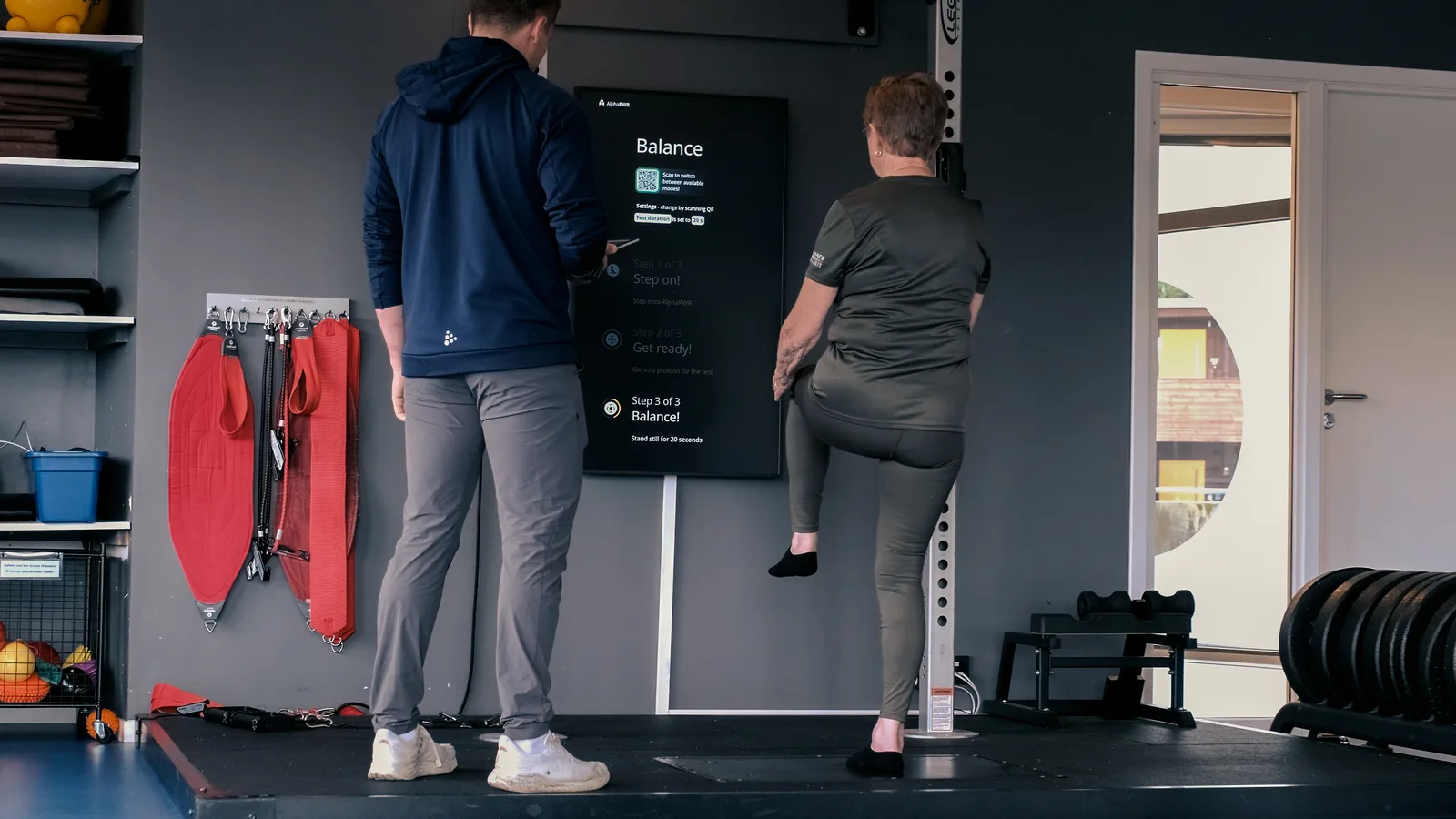 En eldre pasient står på ett bein på Alphatek-plattformen mens skjermen viser instruksjonene for balansetesten.