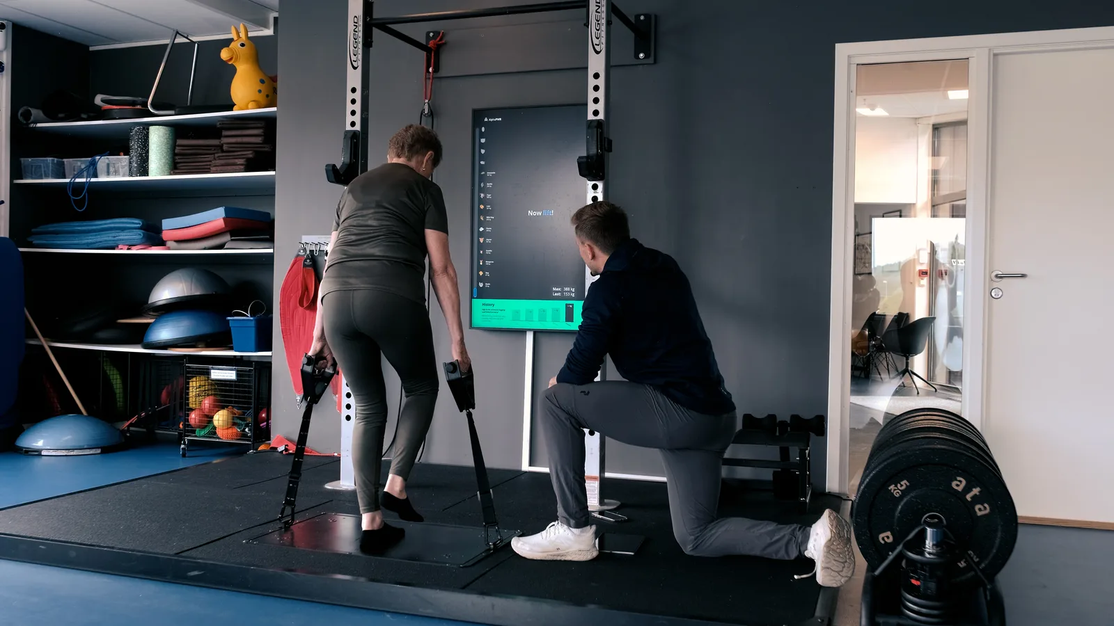 En eldre pasient gjør en øvelse med håndtak på Alphatek-plattformen mens fysioterapeuten følger med ved skjermen.