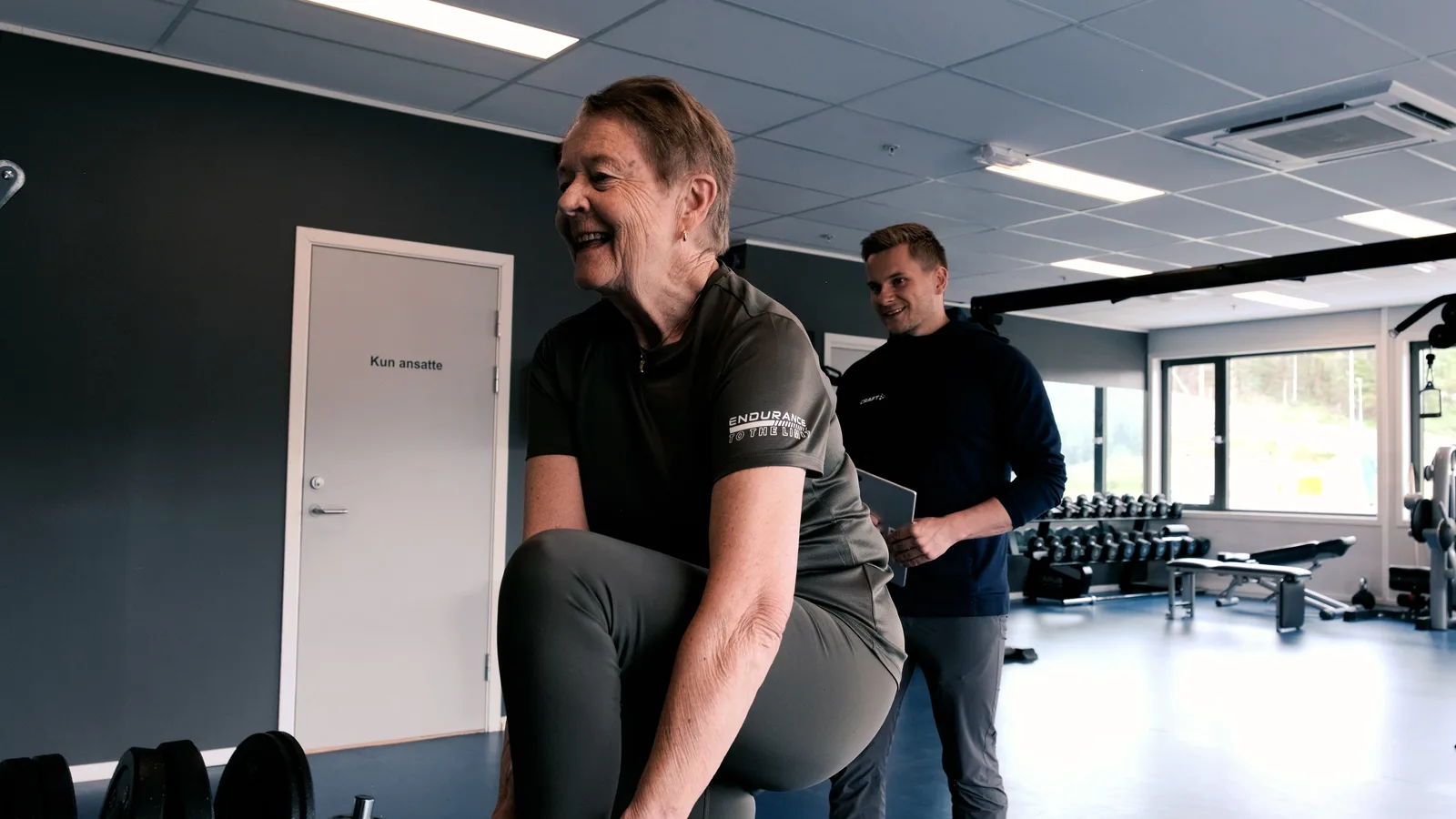 En smilende eldre pasient gjør en øvelse mens fysioterapeuten følger med med et nettbrett.