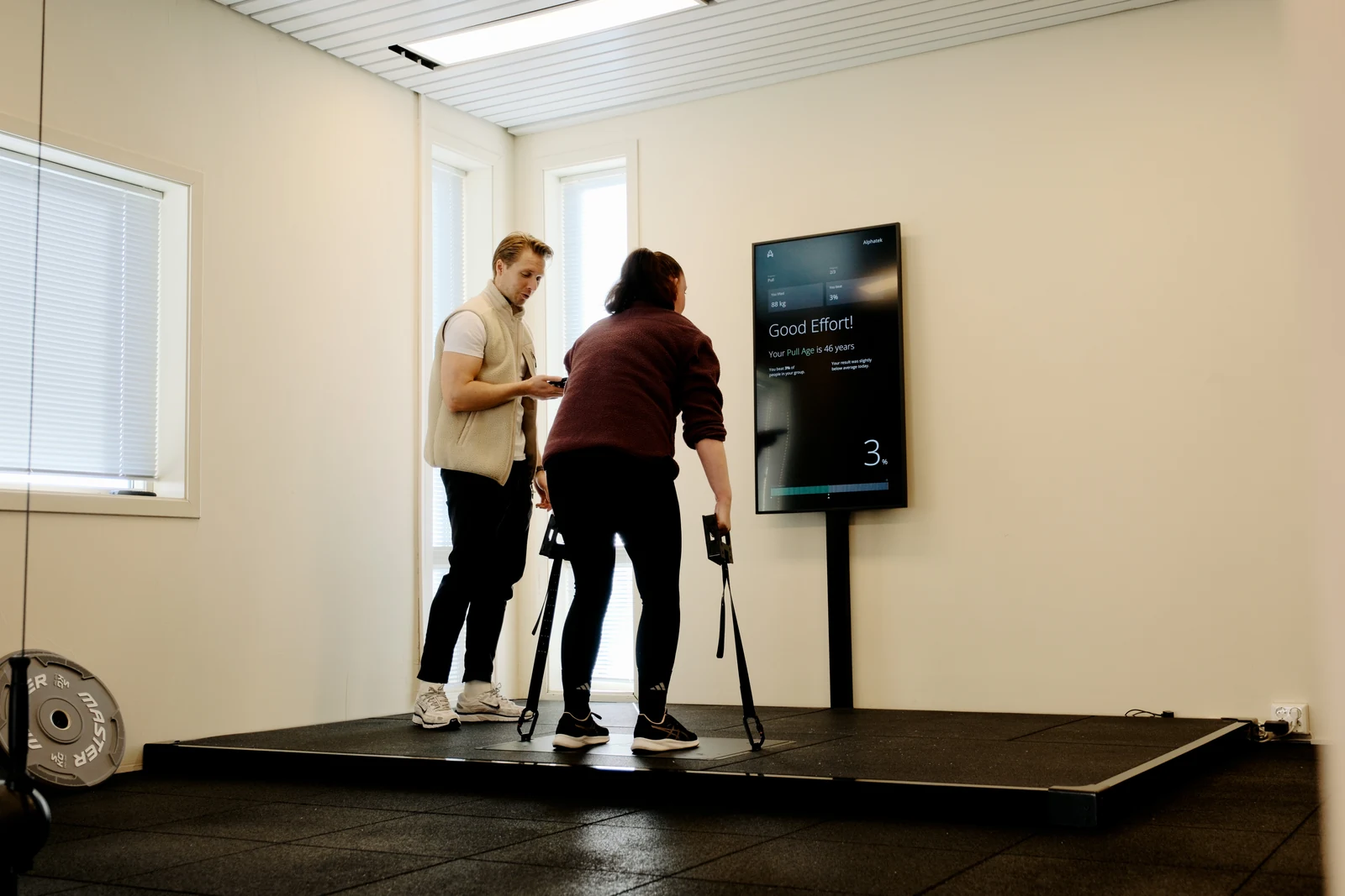 A participant holds handles on the Alphatek platform while a guide observes and the display shows a Fitness Age result.