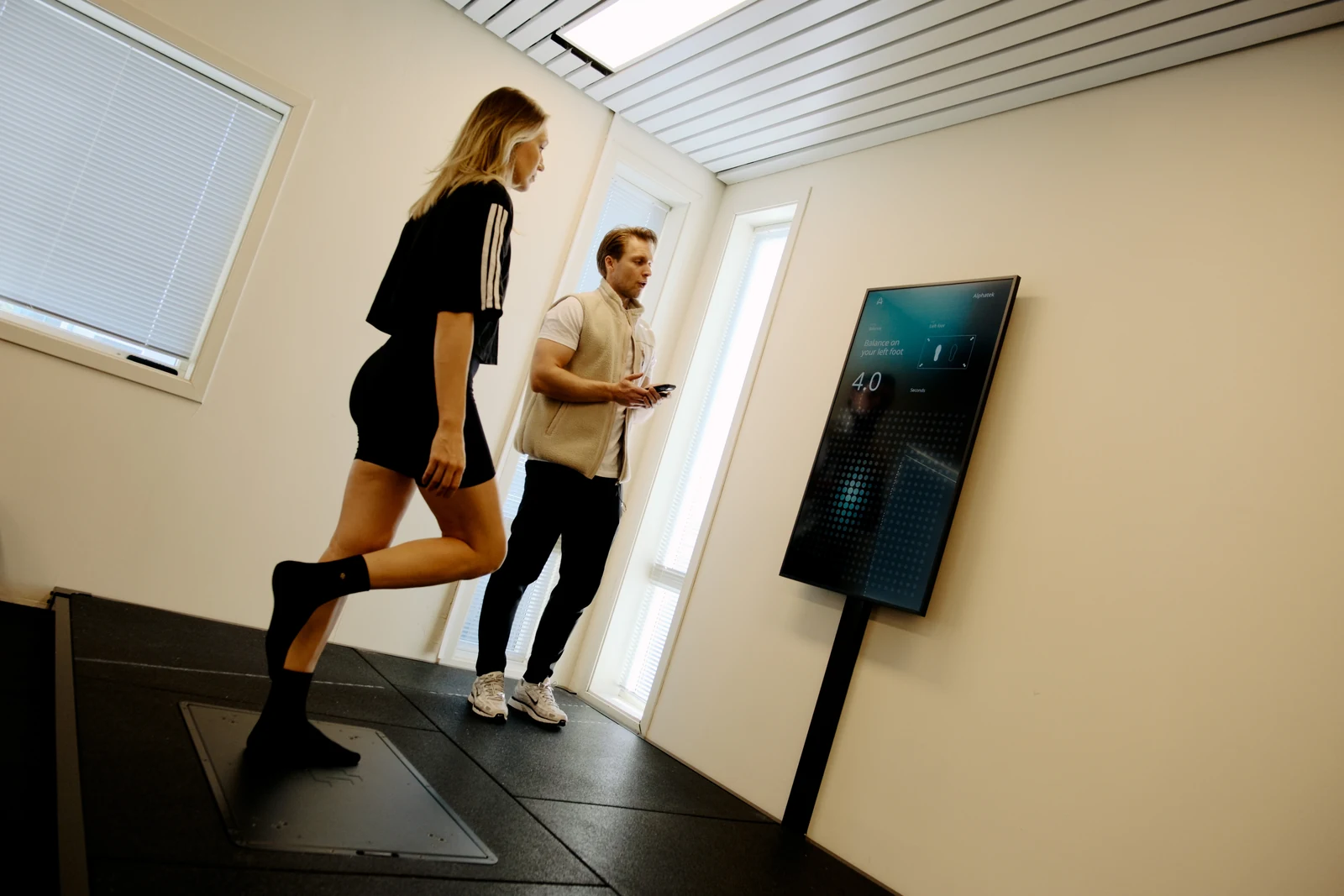Et medlem står på ett bein på Alphatek-plattformen mens treneren veileder ved skjermen.