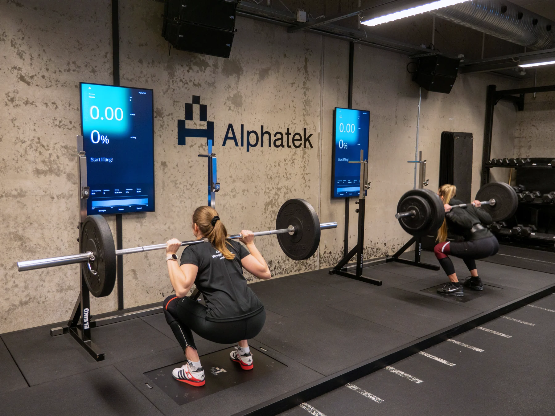 Two people train squats on separate Alphatek stations with large displays in front of them.