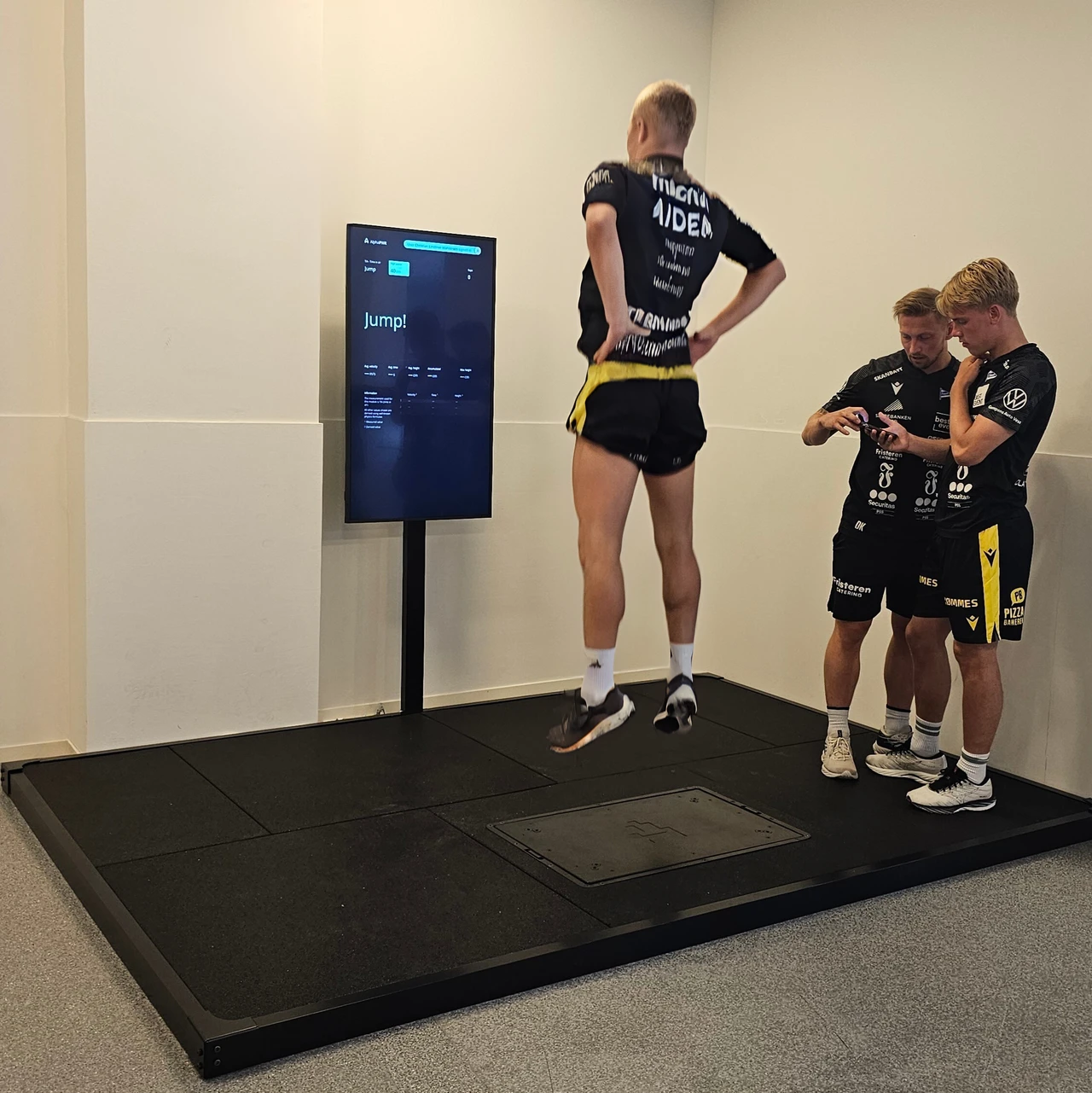 En utøver hopper på Alphatek-plattformen mens to andre følger med på en telefon ved siden av.