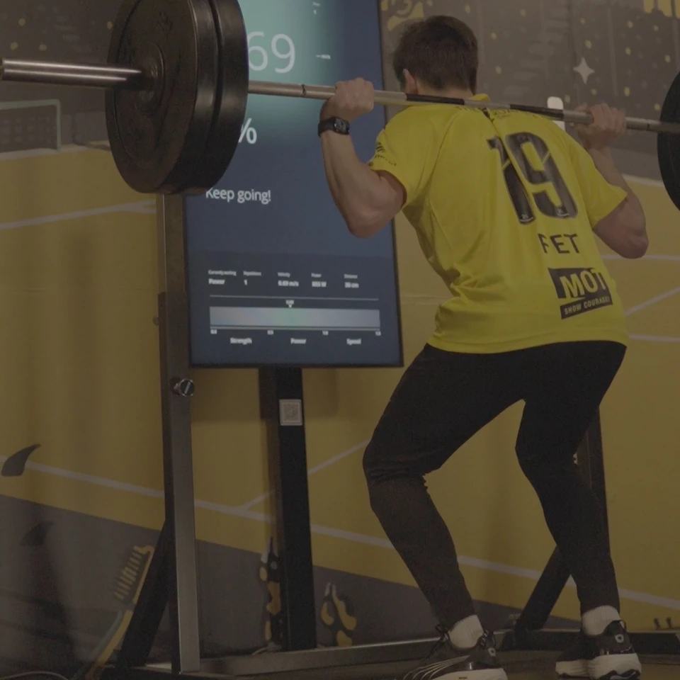 En utøver i gul Bodø/Glimt-drakt utfører knebøy med vektstang foran Alphatek-skjermen.